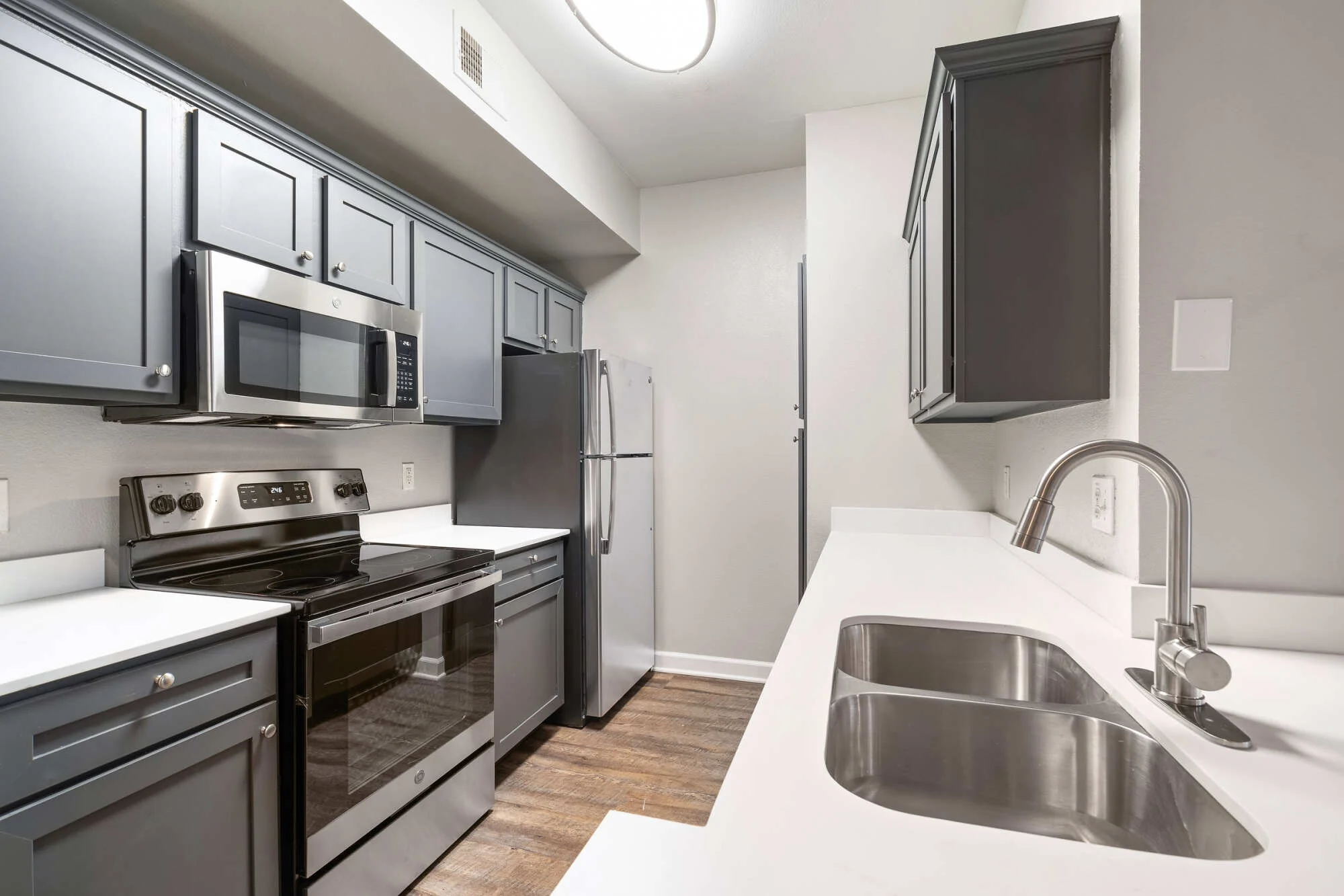 Galley apartment kitchen with gray shaker cabinets, white quartz countertops, stainless steel appliances, a deep double sink, and warm wood-look flooring.